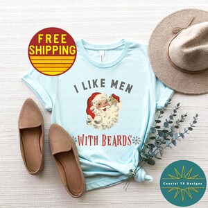 Op de afbeelding: Lichtblauw T-shirt met een vintage illustratie van de kerstman die een cadeau vasthoudt. De tekst "Ik hou van mannen met baarden" staat op het T-shirt gedrukt.