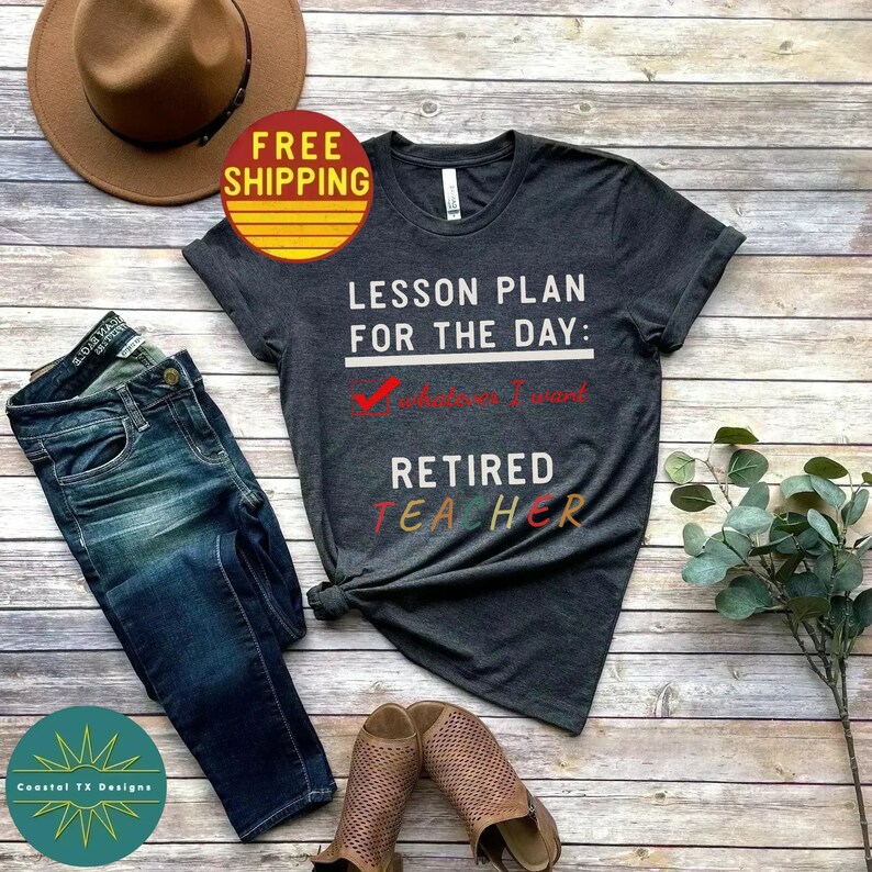 Op de afbeelding: Een donkergrijs T-shirt met de tekst "Lesson Plan for the Day: Whatever I Want Retired Teacher" in witte en kleurrijke letters. Het shirt ligt op een houten achtergrond met een paar bruine sandalen, een blauwe jeans en een bruine hoed.