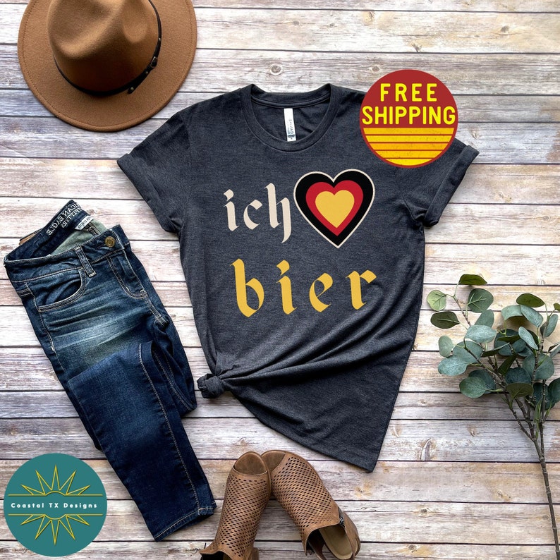 May include: A dark gray t-shirt with the text "ich &hearts; bier" in yellow and black lettering. The heart is red, yellow, and black. The shirt is on a wood background with a hat, jeans, and sandals.