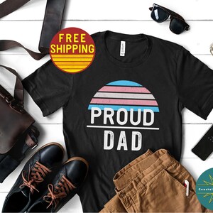 K&ouml;nnte beinhalten: Schwarzes T-Shirt mit einem Sonnenuntergangs-Motiv in den Farben der Transgender-Pride und dem Text "Proud Dad".