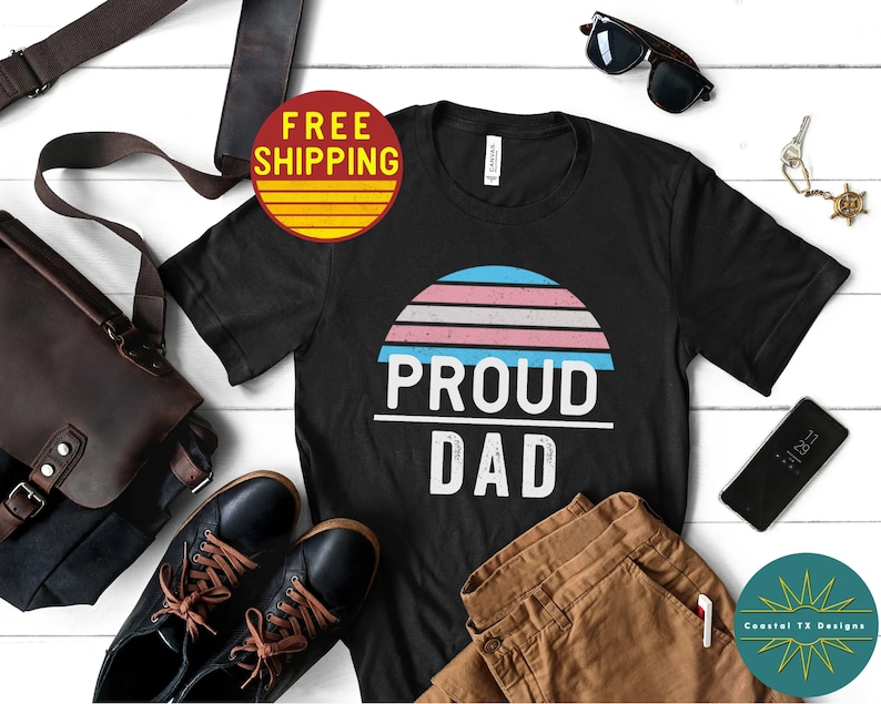 K&ouml;nnte beinhalten: Schwarzes T-Shirt mit einem Sonnenuntergangs-Motiv in den Farben der Transgender-Pride und dem Text "Proud Dad".
