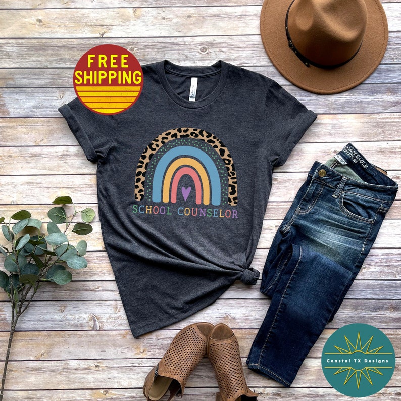 May include: A dark gray t-shirt with a rainbow and leopard print design. The rainbow has a heart in the center and the text "SCHOOL COUNSELOR" is printed below it.