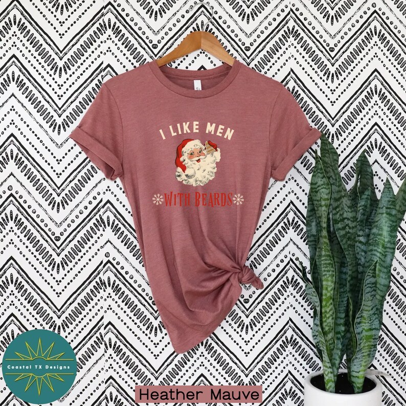 Op de afbeelding: Een heide-paarse t-shirt met een vintage-illustratie van de kerstman die een biertje vasthoudt en de tekst "Ik hou van mannen met baarden".