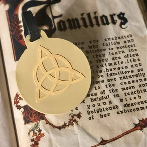 May include: A gold-colored metal charm with a Celtic knot design. The charm is attached to a black leather strap. The charm is sitting on a page of a book with the word "Familiars" printed in black.