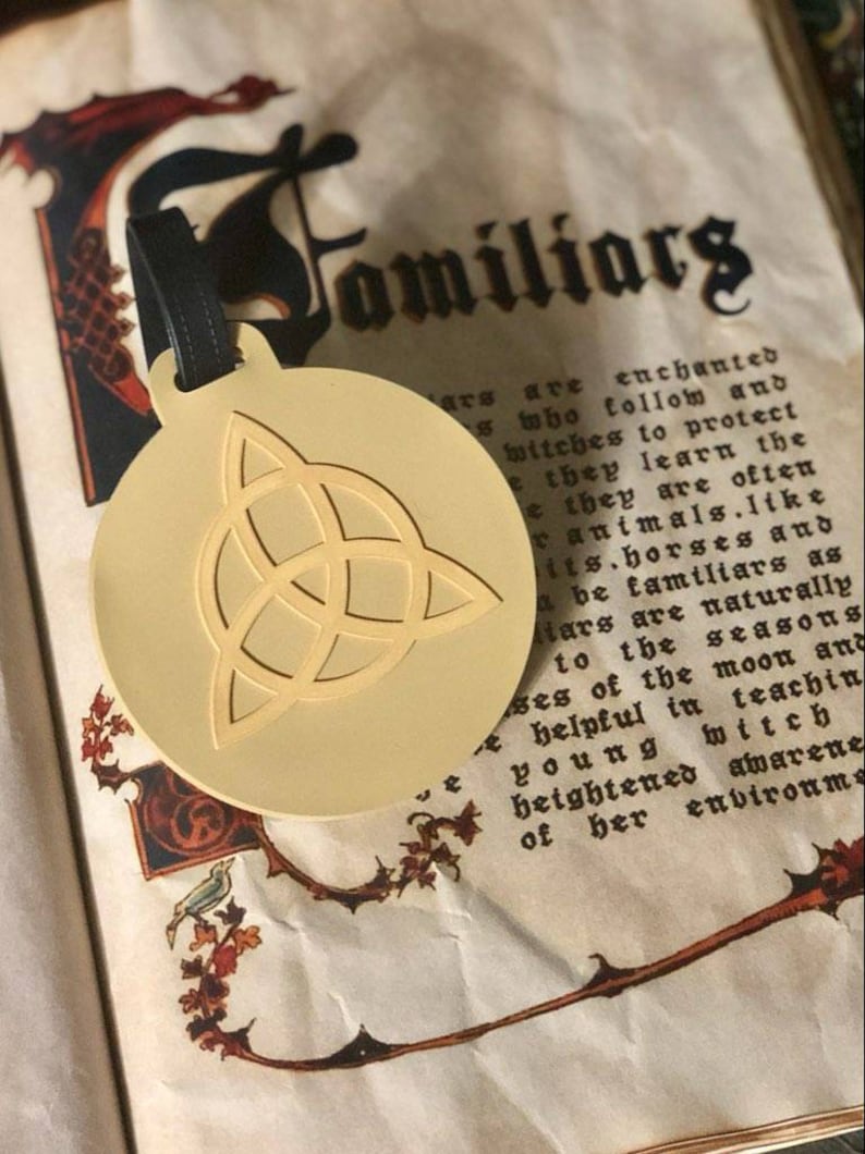 May include: A gold-colored metal charm with a Celtic knot design. The charm is attached to a black leather strap. The charm is sitting on a page of a book with the word "Familiars" printed in black.