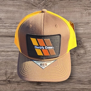May include: Brown and yellow trucker hat with a black patch that says "Herb's Crew" in orange and yellow letters. The patch has a striped design in orange, yellow, and brown. The hat has a brown brim and a yellow mesh back. The hat has a label that says "Premium Quality Headwear 815 Kamel."