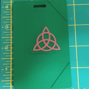 May include: A green plastic badge holder with a pink Celtic knot design. The badge holder has a hole at the top for a lanyard.