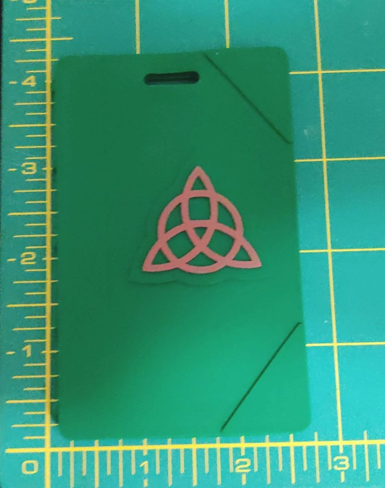 May include: A green plastic badge holder with a pink Celtic knot design. The badge holder has a hole at the top for a lanyard.