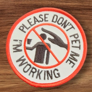 May include: A white embroidered patch with a red circle and a diagonal red line through it. The text "I'm Working Please Don't Pet Me" is written in a gray, cursive font around the circle. Inside the circle is a gray silhouette of a person with a hand reaching towards them.