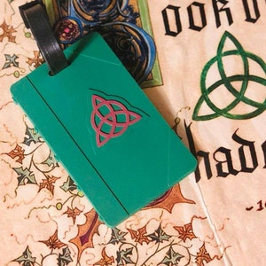 May include: A green luggage tag with a pink triquetra symbol. The tag is attached to a black luggage strap. The tag is lying on a cream-colored surface with a green and blue design.