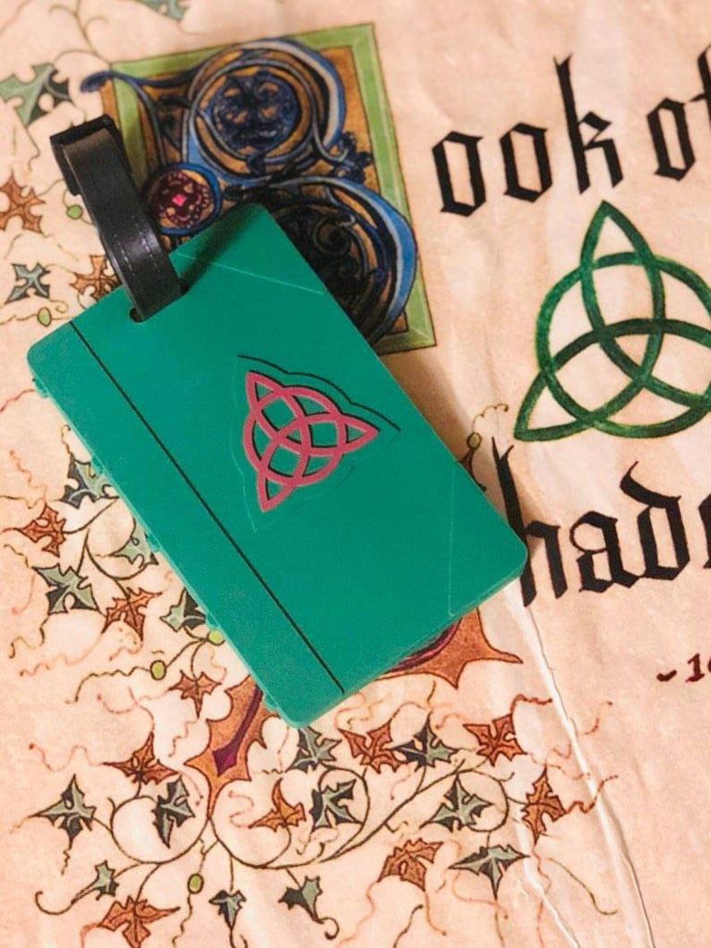 May include: A green luggage tag with a pink triquetra symbol. The tag is attached to a black luggage strap. The tag is lying on a cream-colored surface with a green and blue design.
