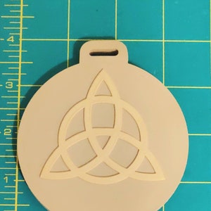 May include: A beige round ornament with a white triquetra symbol. The ornament has a hole at the top for hanging.