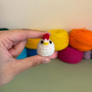 May include: A small, round, white crocheted chicken amigurumi featuring a bright red comb on top, a yellow beak, and two small black bead eyes. The handcrafted yarn figure is spherical in shape and displays a textured, knotted stitch pattern.