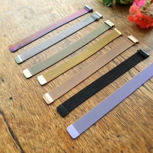 May include: Seven different coloured metal watch straps laid out on a wooden surface. The straps are in the following order from left to right: iridescent, grey, khaki, gold, rose gold, black, and lavender.