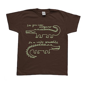 May include: Brown t-shirt with a light green alligator design and the phrases "See you later alligator" and "In a while crocodile" in a handwritten style. The shirt has a crew neck and short sleeves.