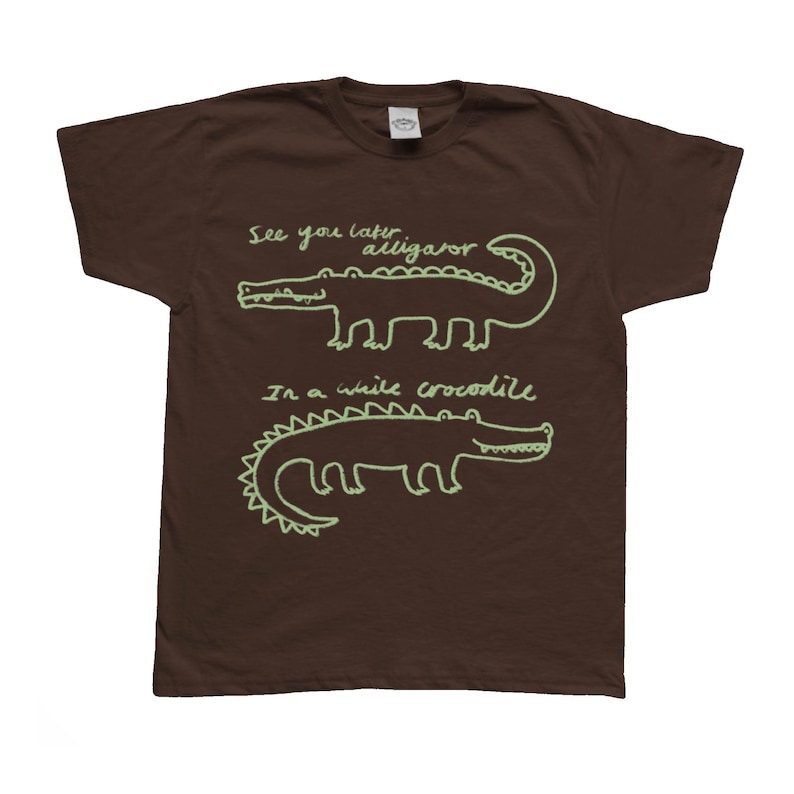 May include: Brown t-shirt with a light green alligator design and the phrases "See you later alligator" and "In a while crocodile" in a handwritten style. The shirt has a crew neck and short sleeves.