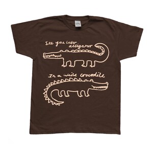 May include: A brown t-shirt with white text and illustrations of two alligators. The top alligator has the text "See you later alligator" above it. The bottom alligator has the text "In a while crocodile" below it.