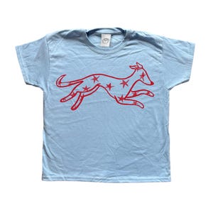 May include: A light blue t-shirt featuring a red graphic of a running dog with star-shaped cutouts. The shirt has short sleeves and a crew neck. The graphic is centered on the front of the shirt.