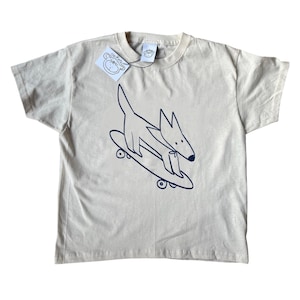 May include: A cream-coloured t-shirt with a navy blue illustration of a dog skateboarding. The dog is depicted with a long tail and pointed ears. The shirt has a tag with a monkey logo.