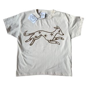 May include: A light beige t-shirt with a brown running dog design filled with stars. The shirt has a crew neck and short sleeves. A small tag with a cartoon monkey logo is attached to the collar.