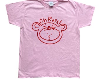 Può includere: T-shirt rosa chiaro con un disegno di ratto rosso a fumetti e la scritta "Oh Rats!". La maglietta ha un girocollo e sembra essere di cotone. Il design è semplice e audace.