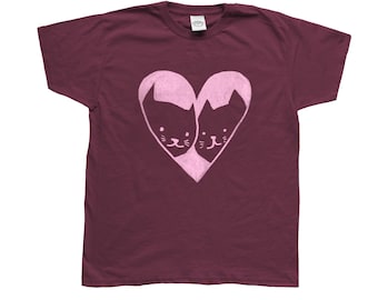Può includere: T-shirt bordeaux con un design a forma di cuore rosa chiaro contenente due sagome di gatti. I volti dei gatti sono visibili all'interno del cuore, con dettagli come occhi, nasi e baffi. La maglietta è realizzata in materiale morbido.