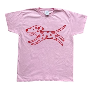 May include: A light pink t-shirt featuring a red cartoon dog with spots, mid-stride. The dog's design is simple and playful, with a smiling face and a curved tail. The shirt is made of soft material.