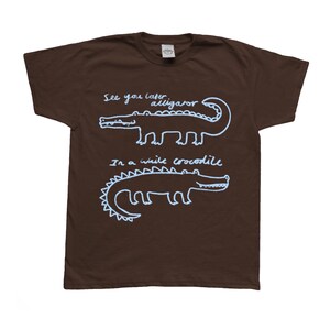 May include: A brown t-shirt with white text and illustrations of alligators and crocodiles. The text reads "See you later alligator" and "In a while crocodile".