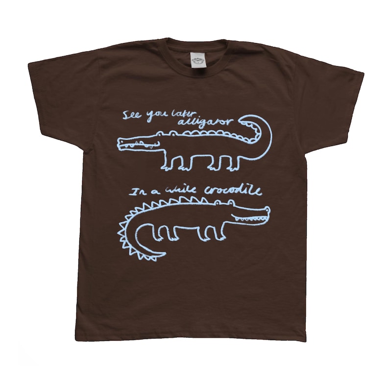 May include: A brown t-shirt with white text and illustrations of alligators and crocodiles. The text reads "See you later alligator" and "In a while crocodile".
