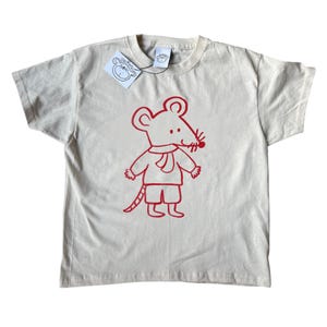 May include: Off-white t-shirt with a red cartoon mouse design. The mouse wears a scarf and shorts. The shirt has a tag with a mouse illustration and the text "On Kate".