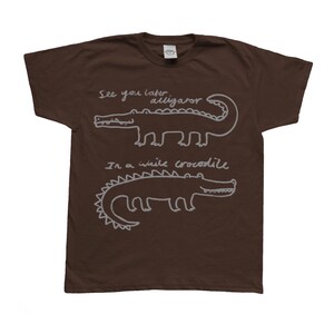 May include: A brown t-shirt featuring two white cartoon alligators with the phrases "See you later alligator" and "In a while crocodile" written above and below the alligators. The shirt is made of a soft material.