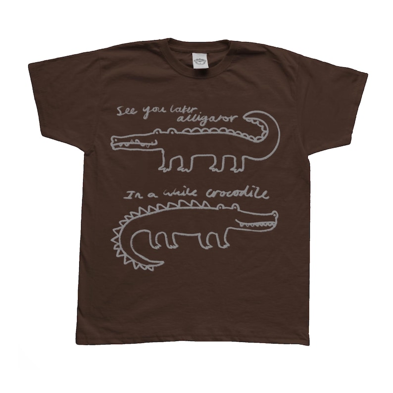 May include: A brown t-shirt featuring two white cartoon alligators with the phrases "See you later alligator" and "In a while crocodile" written above and below the alligators. The shirt is made of a soft material.