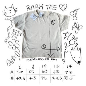 May include: Cream-colored baby tee with measurements in centimeters. The text "BABY TEE" is at the top. The shirt has a tag and is surrounded by whimsical doodles of a cat, worm, bear, flower, snail, cherries, butterfly, star, and dog.