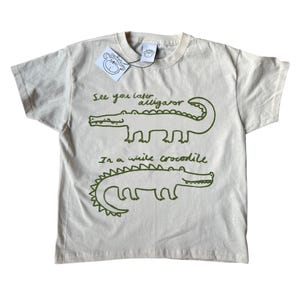 May include: A beige t-shirt featuring green alligator drawings and the phrase "See you later, alligator, in a while crocodile." The top has a crew neck and short sleeves. A tag with a monkey illustration is attached.