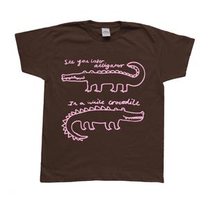 May include: A brown t-shirt featuring two pink alligator/crocodile illustrations with the phrases "See you later alligator" and "In a while crocodile" written above and below the illustrations.