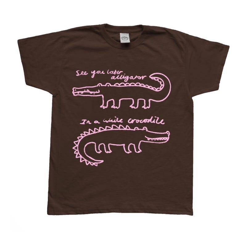 May include: A brown t-shirt featuring two pink alligator/crocodile illustrations with the phrases "See you later alligator" and "In a while crocodile" written above and below the illustrations.