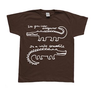 May include: A brown t-shirt with white text and illustrations. The shirt features two alligator/crocodile drawings with the phrases "See you later alligator" and "In a while crocodile". The design is simple and playful.