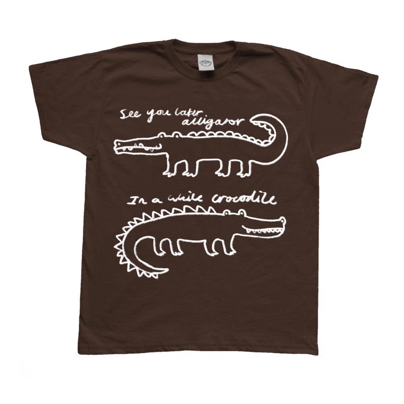 May include: A brown t-shirt with white text and illustrations. The shirt features two alligator/crocodile drawings with the phrases "See you later alligator" and "In a while crocodile". The design is simple and playful.