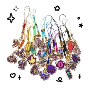 May include: A collection of colorful acrylic charms with various designs, including a hammer, shield, and abstract shapes. Each charm is attached to a gold-colored clasp and a matching colored cord. The background is white with black doodles.