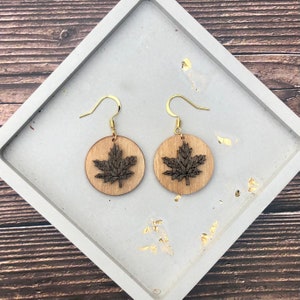May include: A pair of wooden earrings with a maple leaf design. The earrings are brown and have gold hooks.