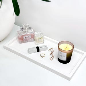 May include: A white rectangular tray displays beauty items. There are perfume bottles, a lipstick, a gold ring, earrings, and a lit candle in a glass jar with the word "CALM" on the label.