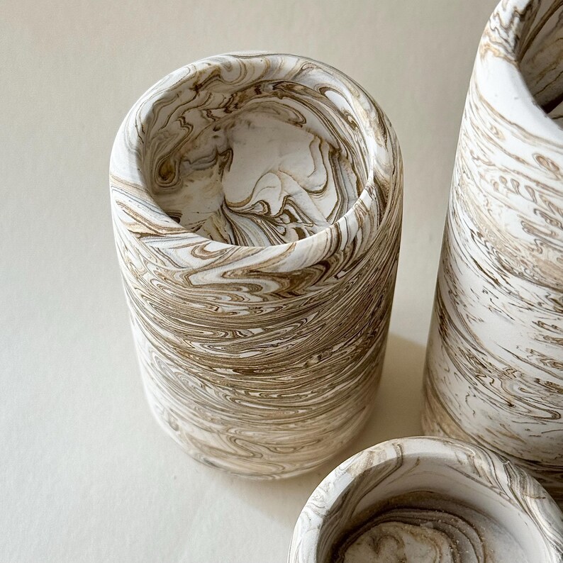May include: A set of cylindrical containers with a marbled design. The containers have a swirling pattern of white and brown tones. The vessels vary in size and shape, with a smooth, polished finish.