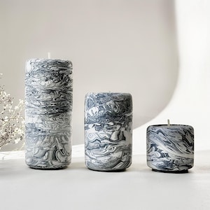May include: Three marbled candles in different sizes. The candles have a swirling pattern of grey and white, similar to stone. Each candle has a wick at the top. The candles are cylindrical and sit on a white surface.
