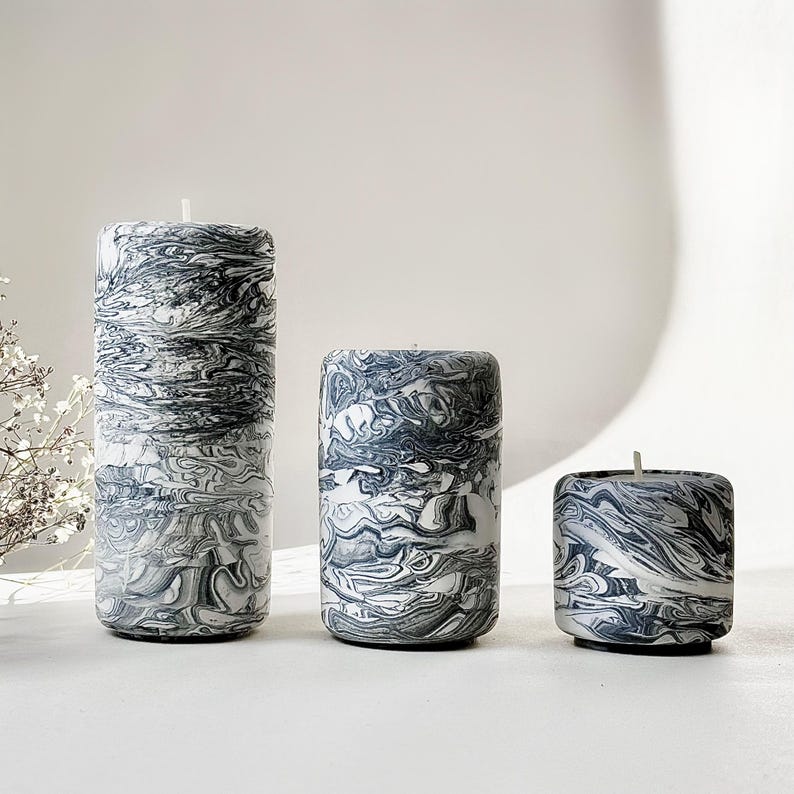 May include: Three marbled candles in different sizes. The candles have a swirling pattern of grey and white, similar to stone. Each candle has a wick at the top. The candles are cylindrical and sit on a white surface.