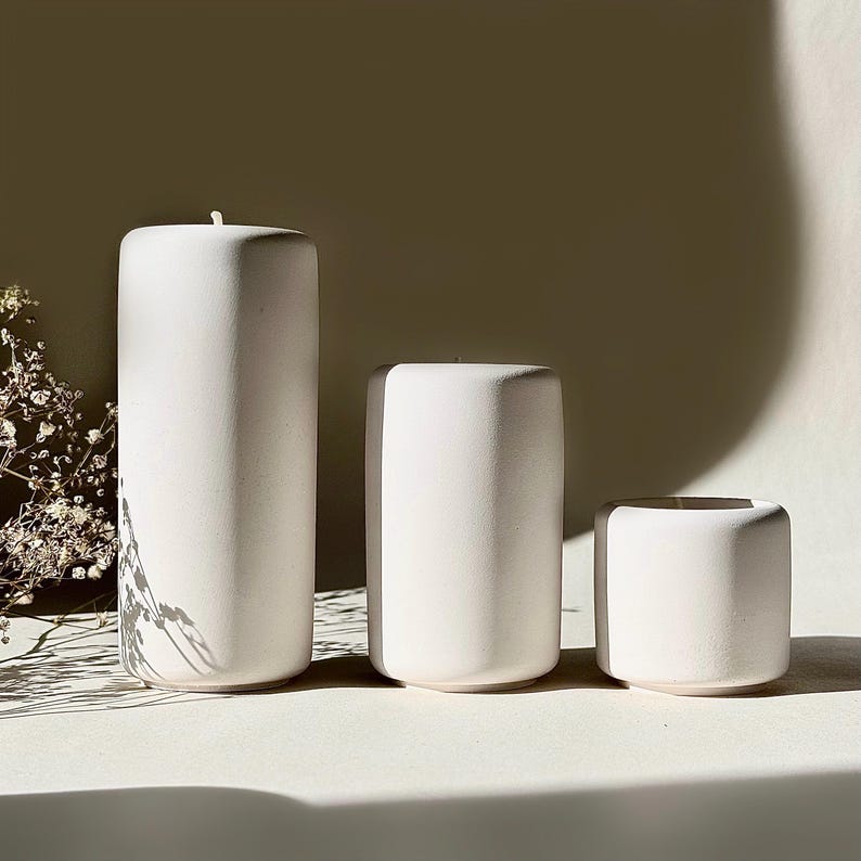 May include: Three white, cylindrical candles of different heights. The tallest candle has a wick. The candles are arranged in a row, with the smallest candle on the right. The background is a neutral colour.