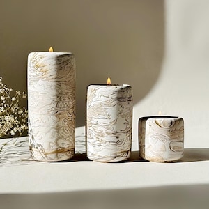 May include: Three lit pillar candles with a white and gold marbled pattern. The candles are of varying heights, with the tallest on the left. The flames are yellow, and the overall style is elegant and sophisticated.