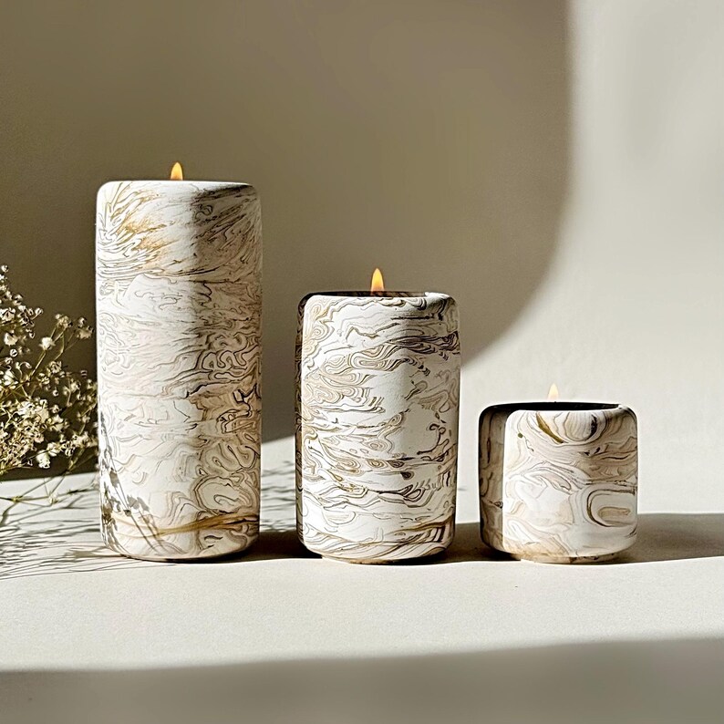 May include: Three lit pillar candles with a white and gold marbled pattern. The candles are of varying heights, with the tallest on the left. The flames are yellow, and the overall style is elegant and sophisticated.