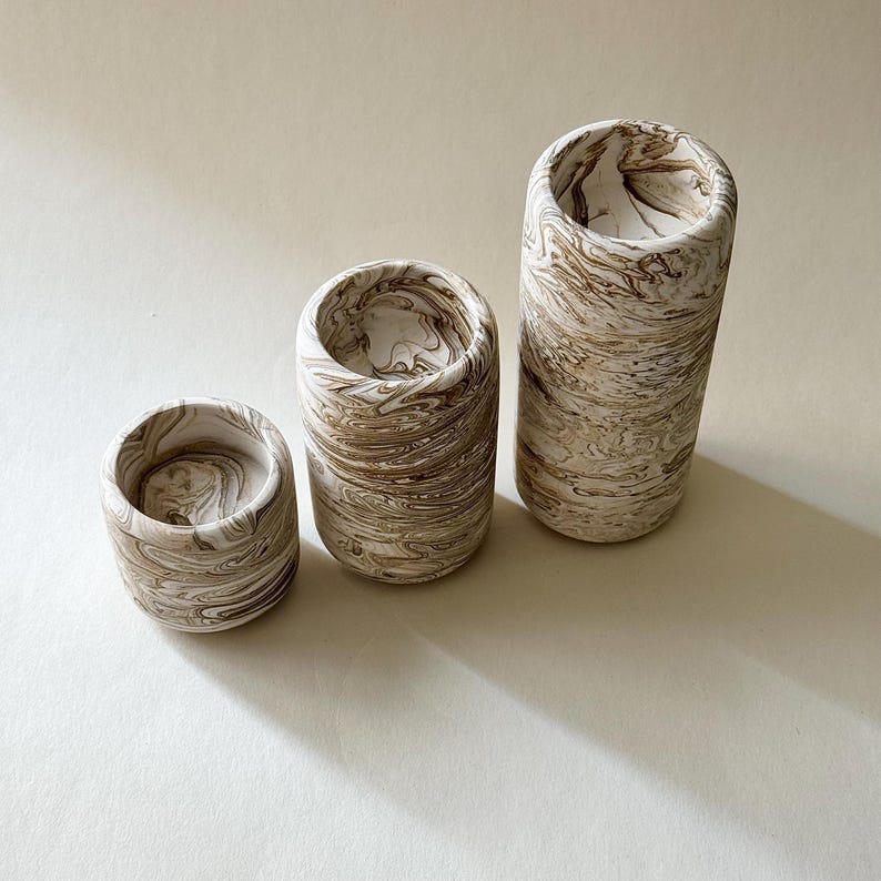 May include: Three cylindrical decorative vases in varying heights. The vases have a marbled pattern in shades of beige and brown. The vases are arranged from smallest to largest, with open tops and rounded bases. The background is a solid, neutral colour.