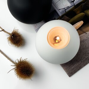 Op de afbeelding: Een ronde witte keramische kaarshouder met een brede opening. De kaarshouder heeft een brandende kaars erin, waardoor een warm gloed ontstaat. De kaarshouder staat op een tijdschrift met de tekst "Bespoke Sofa" erop gedrukt.