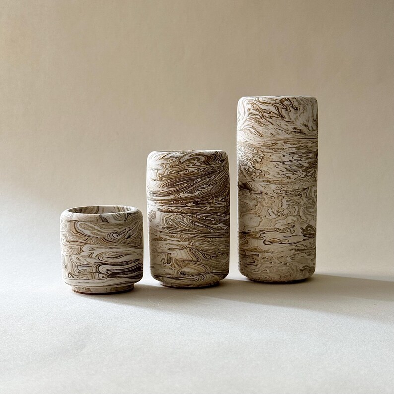 May include: Three cylindrical ceramic vases with a marbled pattern in shades of brown and cream. The vases vary in height, with the tallest being approximately 25 cm tall. The design features swirling lines, creating a unique texture.